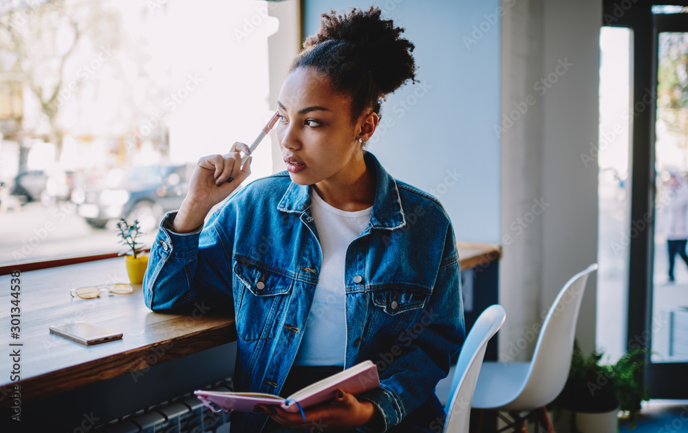 Thoughtful female blogger looking away and feeling pondering on ideas for new article at own web page, pensive woman sitting in cafeteria with textbook in hand and thinking about plan for future trip - obrazy, fototapety, plakaty 