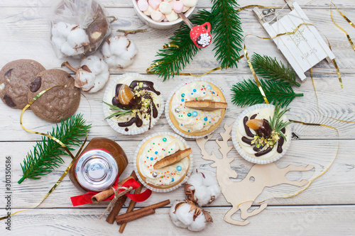 Cupcakes with cream cheese frosting and tangerines, cotton flowers, christmas tree branches, mini wooden sledge, chocolate cookies, caramel jar with red riboon, cinnamon rolls on wooden background. photo