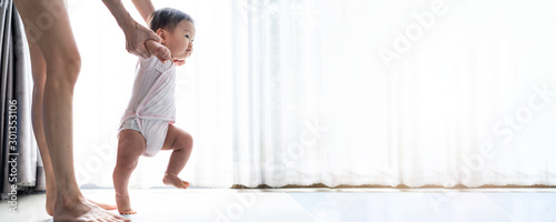 Asian baby taking first steps walk forward on the mat. Happy little baby learning to walk with mother help at home. Mother teaching how to walk gently. Baby growth and development concept. Banner size photo