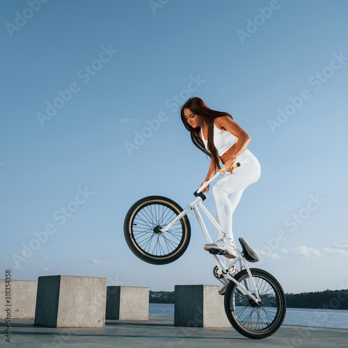 Doing stunts. Female rider is on the bike at daytime near the lake. Fitness woman in sportive clothes