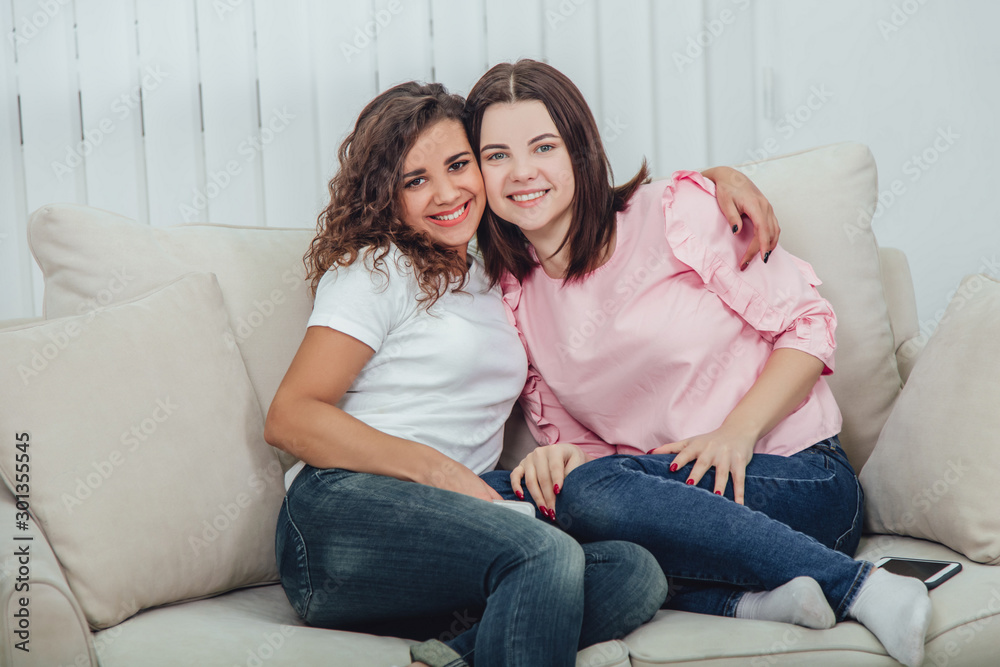 Two amazing girls sitting on the sofa, hugging one another, smiling at the camera.