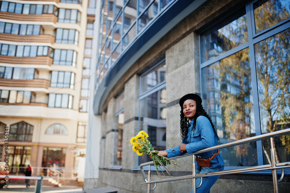 Stylish fashionable african american women in jeans wear and black beret with yellow flowers bouquet posed outdoor in sunny day against blue modern building.