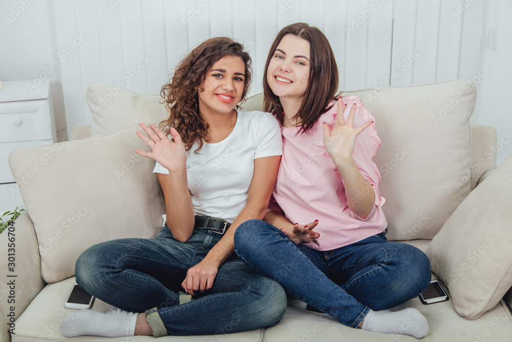 Two amazing girls sitting on the sofa, side by side, in lotos position, waving hello and smiling at the camera.