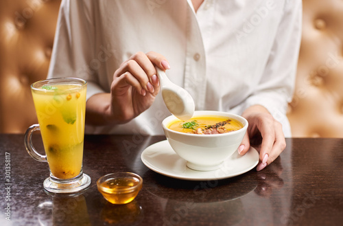 Close up woman in white blouse holding in hands white bowl with orange cream soup. Female enjoying in restaurant with first dish and tea with mint and honey near. Concept of healthy food.