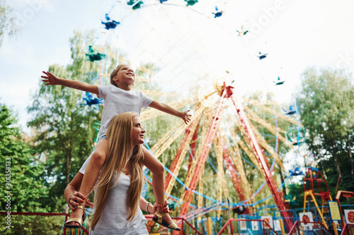 Daughter sits on the shoulders. Cheerful little girl her mother have a good time in the park together near attractions photo