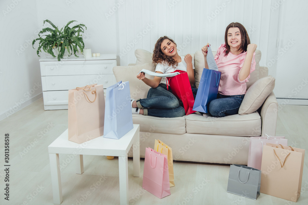 Extremely happy schopahollic girls sitting on the sofa, taking out clothes from colorful shop bags, smiling. Extremely happy schopahollic girls sitting on the sofa, taking out clothes from colorful shop bags, smiling.