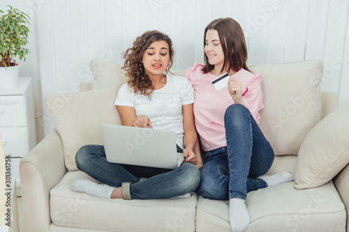 Two happy female friends using laptop for shopping online. One girl is holding credit card. They choose clothes.
