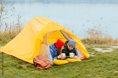 Cute young couple of millennial or generation z adventurers or travellers sit in front of tent, girl makes selfie,man looks into distance contemplating and relaxing. Scandinavian lifestyle inspiration