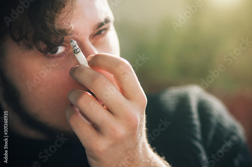 A curly-haired bearded guy in a sweater holds a cigarette near his eye and attentively warily looks with his cunning eyes.
