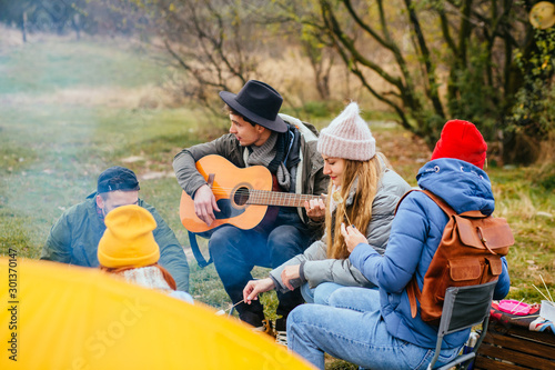 Camping outdoor. Group friends camping leisure and destination travel. Family people sitting around camp fire and playing guitar and roasted sausages, tourism relax and travel near river in holiday.