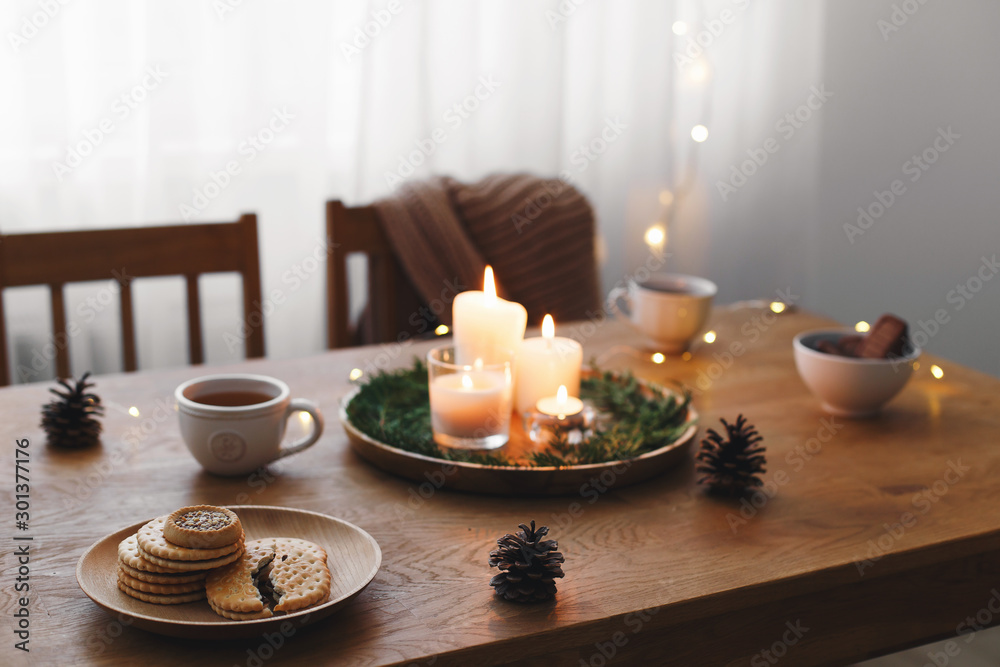 Cozy evening tea party by candlelight. Served table: cup of tea, cookies, tray, candles, pine cone, branches, led garland lights. Table setting. Christmas or New year holiday decorations. Table decor.