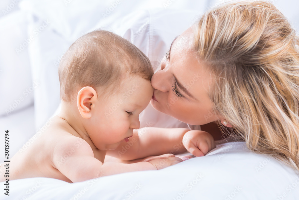 Mother and baby boy son playing on a white bed. Mothers tenderness and kisses of a toddler child Mother and baby boy son playing on a white bed. Mothers tenderness and kisses of a toddler child