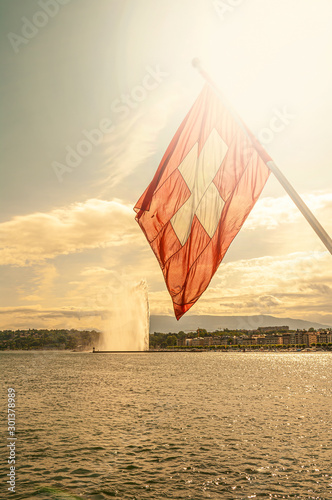 Swiss flag, Leman lake at sunset and a super Jet fountain in the historical downtown of Geneva, Switzerland, summer