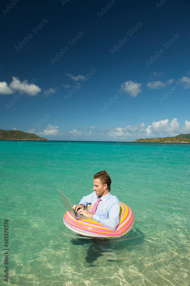 Businessman working on his laptop while floating on a colorful inflatable ring in bright tropical waters
