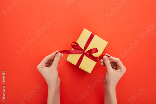 cropped view of woman untying ribbon on golden gift box on red background © LIGHTFIELD STUDIOS