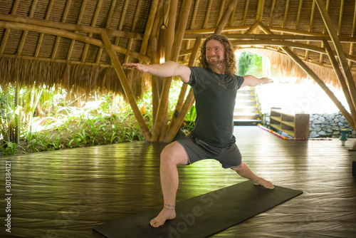 lifestyle portrait of young attractive and happy man in hipster yogi style doing yoga drill on mat at beautiful Asian bamboo hut stretching in mind balance and good vibes