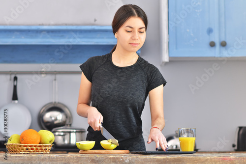 A young girl in a black t-shirt makes a fruit breakfast and cuts apple on a light wooden board in a kitchen.