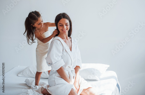 Helping to made hairstyle. Young mother with her daugher have beauty day indoors in white room