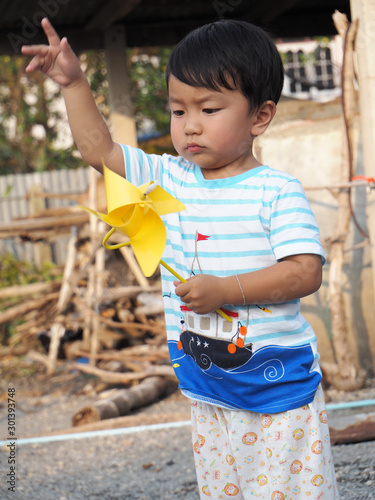 Asian lovely child boy turning yellow windmill toy by hand with countryside background. Cute young kid playing and learning how turbine blow. Freedom and happy time. Preschool learning concept.