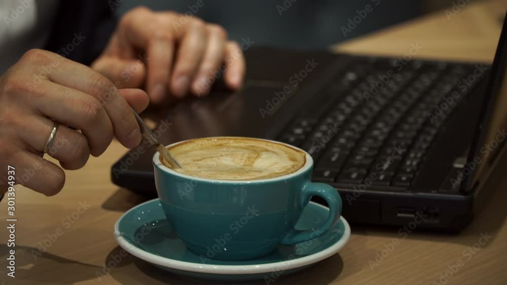 Close up of male hands of businessman in suit stir hot cappuccino coffee with spoon and work typing on keyboard at table in coffee house. Close up of male hands of businessman in suit stir hot cappuccino coffee with spoon and work typing on keyboard at table in coffee house.