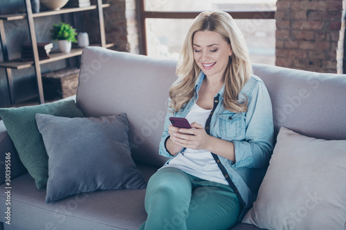 Portrait of her she nice attractive lovely pretty charming cheerful cheery wavy-haired lady sitting on divan using digital device gadget at modern industrial loft brick style interior living-room