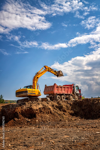 Yellow excavator and empty dump truck working at the construction site. Vertical view
