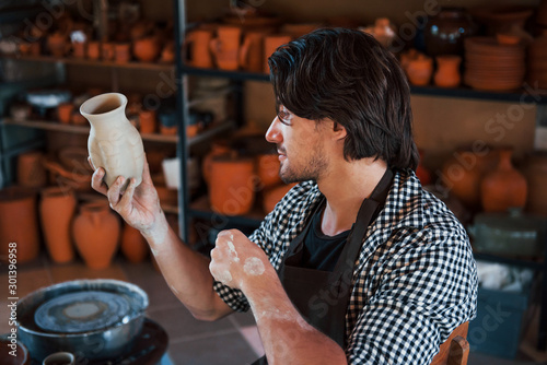 Obraz na plátně Young ceramist holds fresh handmade pot in hand and looks at results of his work