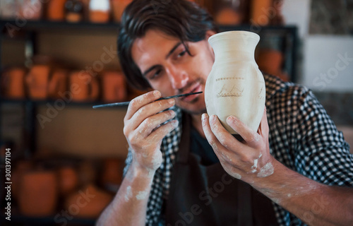 Fotografie Young ceramist holds his fresh handmade pot and makes some detalizing by special
