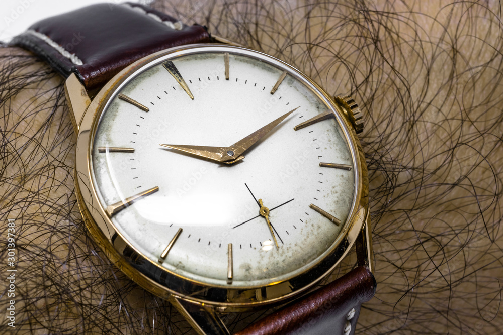 An old gold mechanical wristwatch with manual winding. Antique watch ruined  by time, with scratches and mildew stains. Watch worn on a man's hairy wrist.  Leather band. Vintage clock. Spend time. Photos