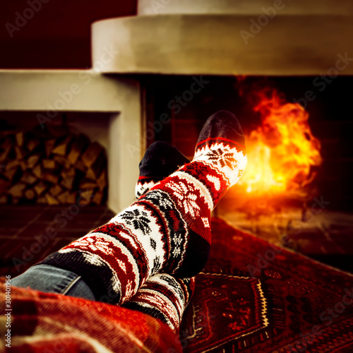 Female legs in winter woolen socks lying on the floor. Fireplace background with burning wood. Red rustic rug on the floor. Christmas. Winter December evening. Copy space. Warm red blanket.