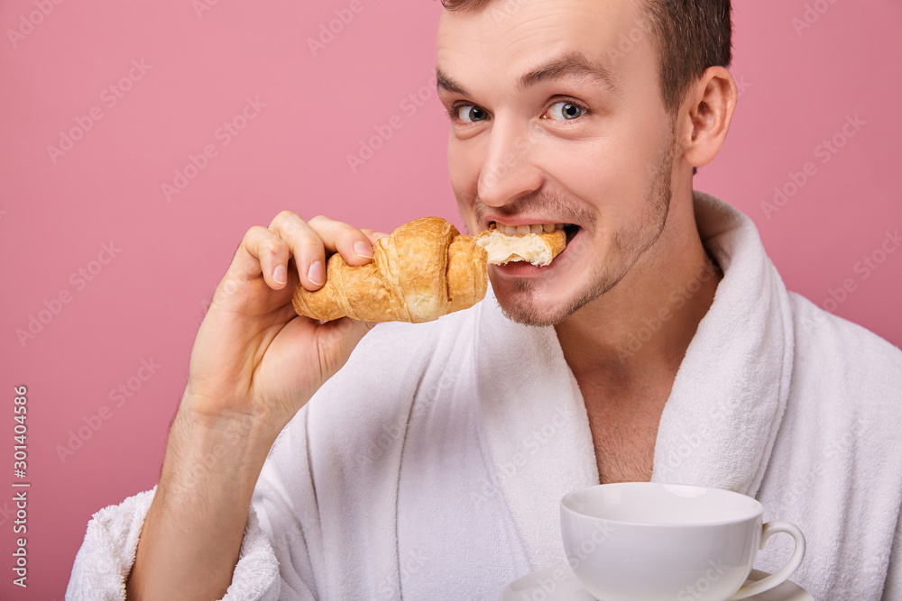 Funny pretty guy is biting fragrant croissant and holding white cup Funny pretty guy is biting fragrant croissant and holding white cup