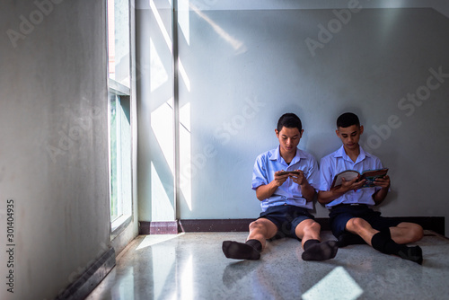 Two male southeast Asian high school students are sitting on the floor. The first student is playing online games. The second student is reading a book.