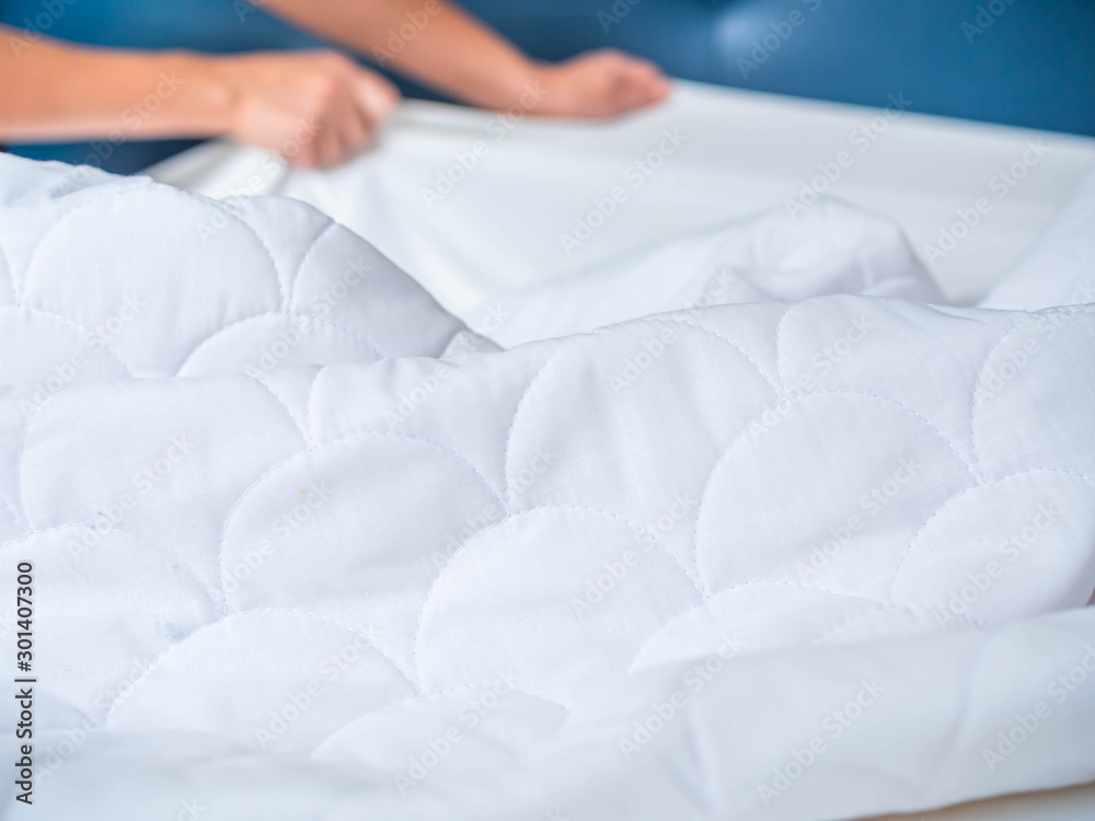 Selective focus at crumpled and wrinkle white bed topper on the bed in the bedroom with blurry woman 's hand pull the bed cover. Housework preparation before laundry process. Selective focus at crumpled and wrinkle white bed topper on the bed in the bedroom with blurry woman 's hand pull the bed cover. Housework preparation before laundry process.