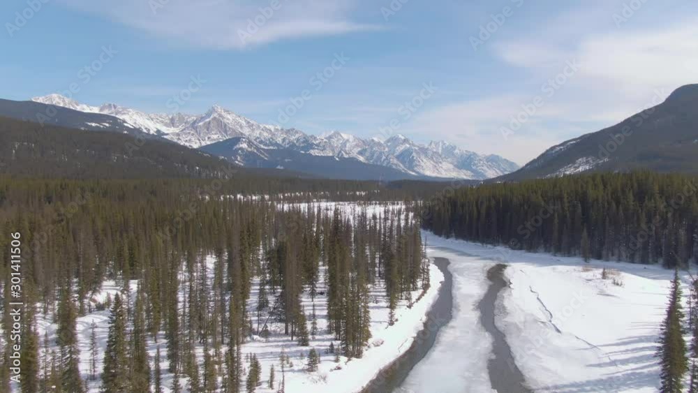 AERIAL: Picturesque shot of river flowing through snowy woods and towards a breathtaking ridge in Alberta. Flying along a mountain stream as it crosses the wintry Canadian wilderness on a sunny day. AERIAL: Picturesque shot of river flowing through snowy woods and towards a breathtaking ridge in Alberta. Flying along a mountain stream as it crosses the wintry Canadian wilderness on a sunny day.