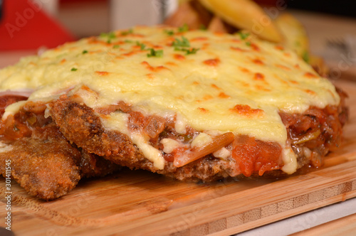 CLASSIC BREADED FRIED MEAT , KNOWN IN ARGENTINA AS MILANESA, CHEESE AND TOMATO, SERVED WITH FRENCH FRIES ON A WOODEN TABLE photo