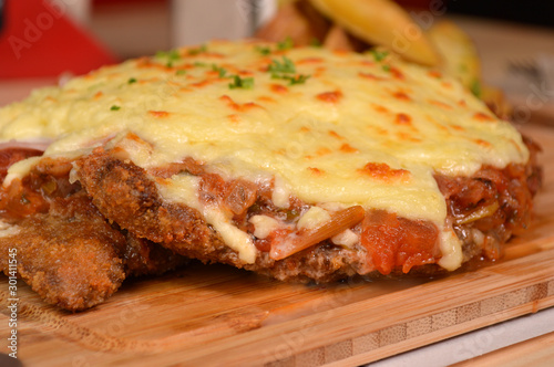 CLASSIC BREADED FRIED MEAT , KNOWN IN ARGENTINA AS MILANESA, CHEESE AND TOMATO, SERVED WITH FRENCH FRIES ON A WOODEN TABLE