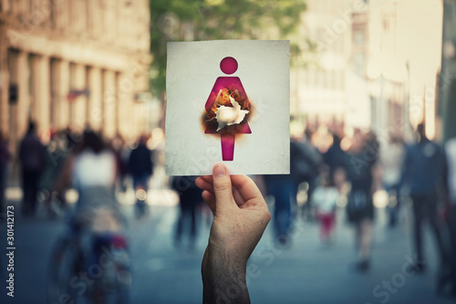 Miscarriage and fetus loss concept as hand holding a paper with woman icon symbol has broken abdomen as a burnt hole in stomach. Human spontaneous abortion message over a crowded street background.