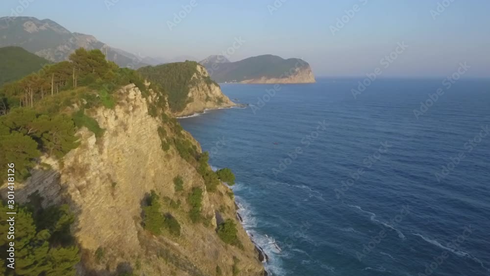 Aerial shot of coastline and sea waves hitting clifs covered with coniferous forest. A small ship sails the sea Aerial shot of coastline and sea waves hitting clifs covered with coniferous forest. A small ship sails the sea
