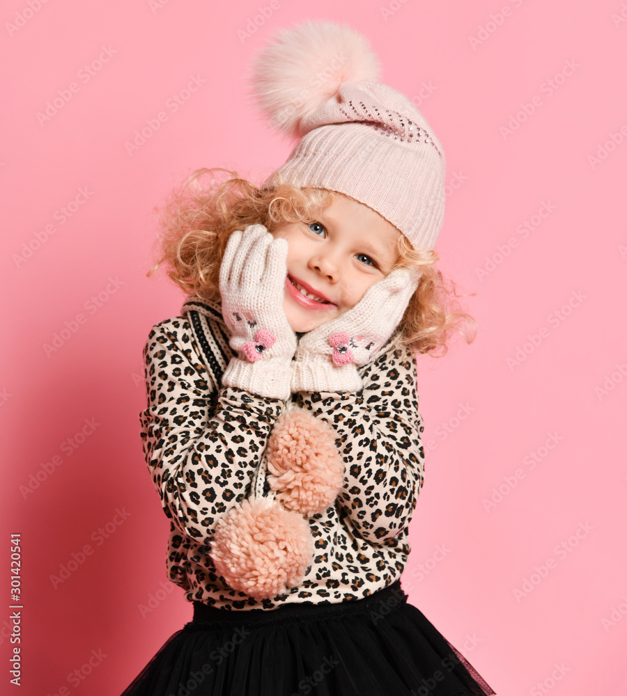 Cute cheerful curly blonde baby girl in knitted hat covering her cheeks with hands in white gloves warming them on pink Cute cheerful curly blonde baby girl in knitted hat covering her cheeks with hands in white gloves warming them on pink
