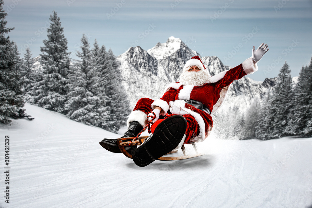 Red Santa Claus riding a wooden sled. An older man with a beard delivers presents to a child. Winter mountains landscape and snow-covered trees with frost. Fir branches, winter time. Christmas spells - obrazy, fototapety, plakaty 