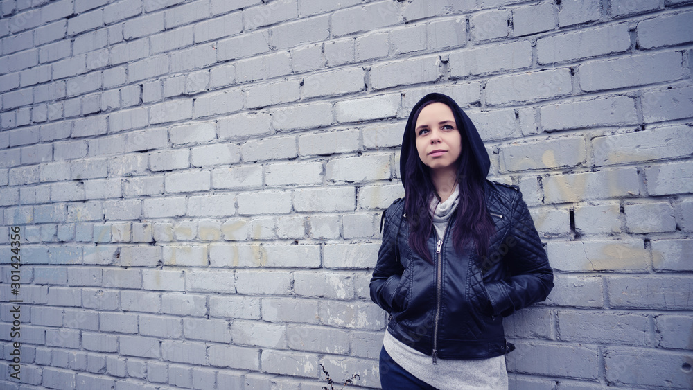 Woman in a black leather jacket. Sad female looking to the side. Young woman in a black leather jacket at the background of a brick wall