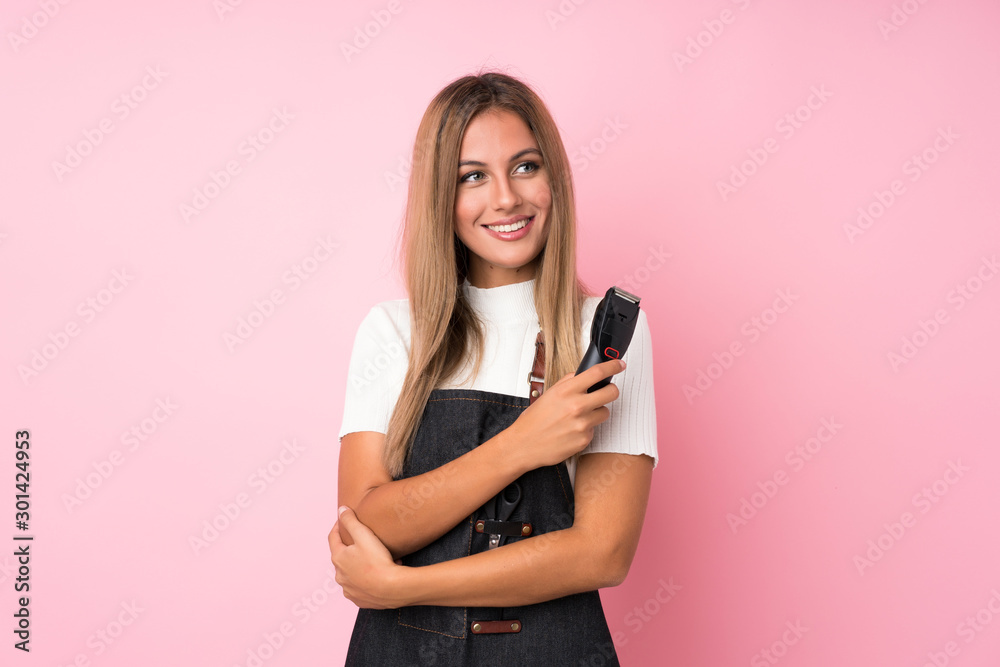 Young blonde woman over isolated pink background with hairdresser or barber dress and holding hair cutting machine Young blonde woman over isolated pink background with hairdresser or barber dress and holding hair cutting machine