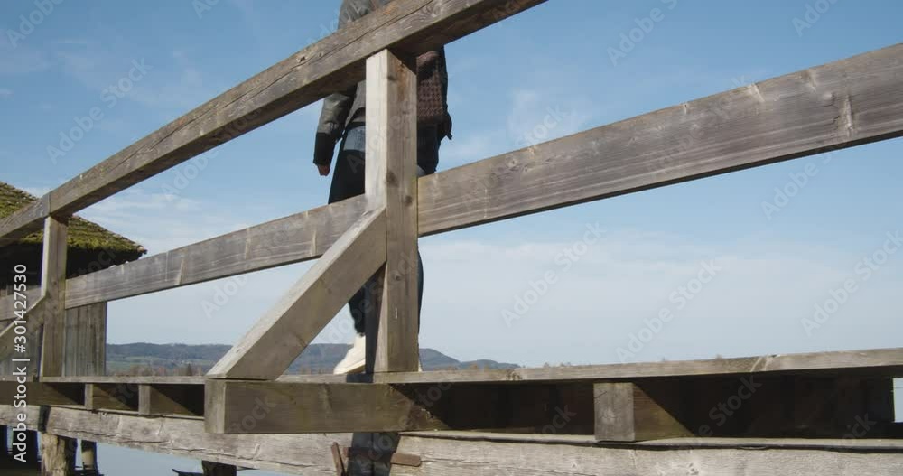 Tracking shot of woman walking down a jetty to a small hut on a reflective clear mountain lake Tracking shot of woman walking down a jetty to a small hut on a reflective clear mountain lake