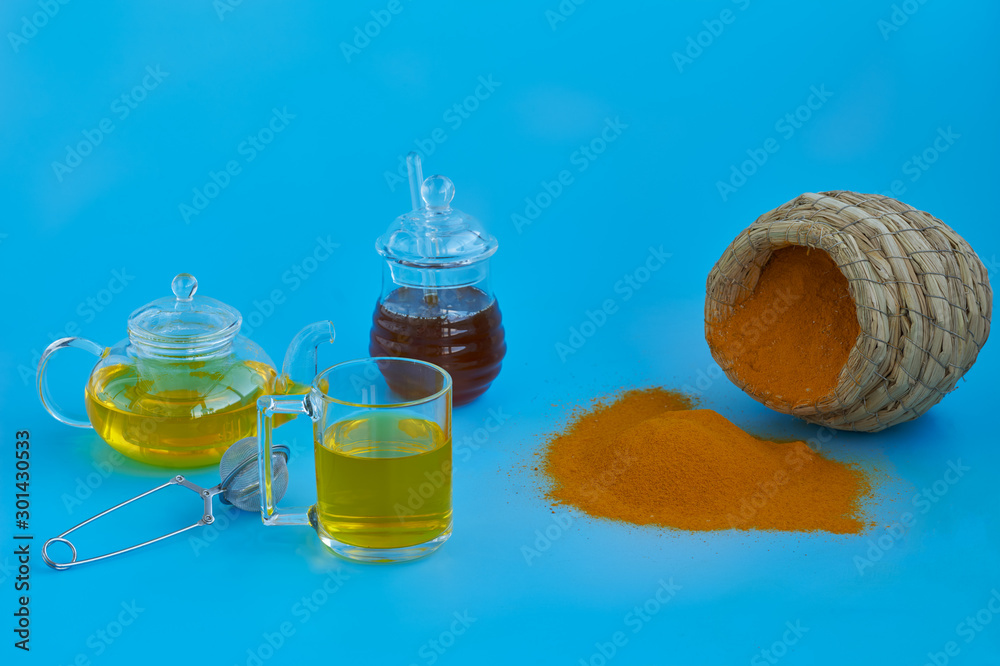 Front view of turmeric powder, a turmeric tea, a teapot, a jar of heather honey and a handmade palm leaf bowl filled with turmeric powder on a blue background. Front view of turmeric powder, a turmeric tea, a teapot, a jar of heather honey and a handmade palm leaf bowl filled with turmeric powder on a blue background.