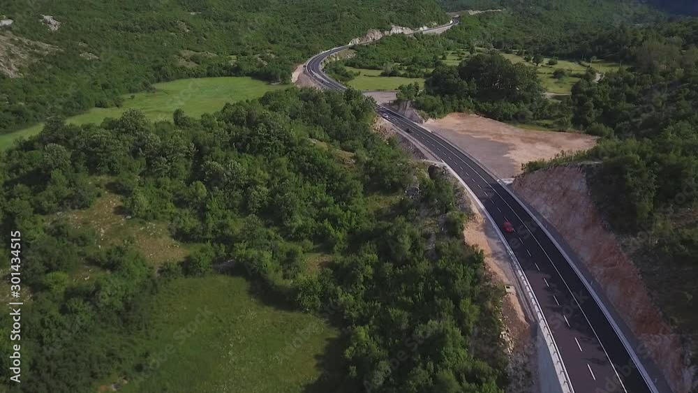 Aerial shot of a newly built highway crossing a mountainous area. The highway is surrounded by hills and green trees and various cars pass through it. Aerial shot of a newly built highway crossing a mountainous area. The highway is surrounded by hills and green trees and various cars pass through it.