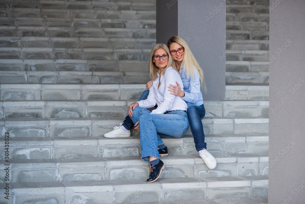 A beautiful blonde with glasses and her mature mother are sitting on the stairs outdoor. A well-groomed elderly woman hugs her adult daughter tightly. Love and family values. Mothers Day.