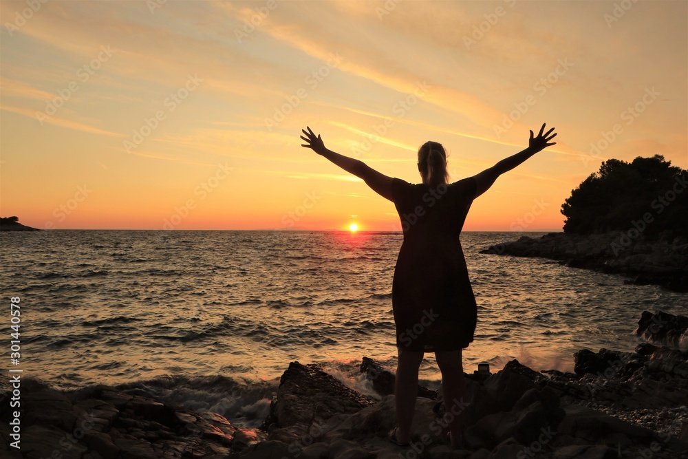 silhouette of a woman standing by the sea with arms raised at sunset