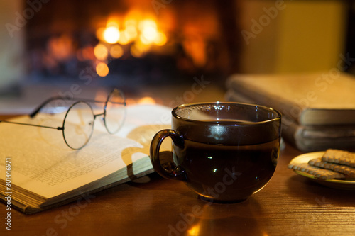 An open ancient book, vintage glasses, a cup and biscuits in front of the fireplace. Evening fairy tales. Fantasy.
