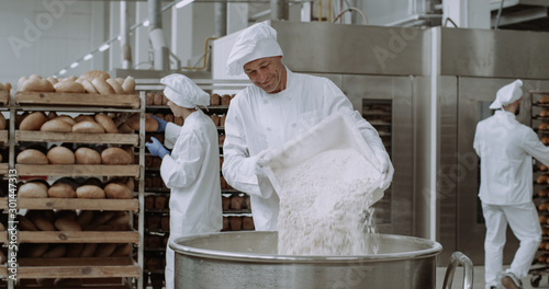 Big bakery industry baker prepare the dough add the flour in a big container background workers arrange the bread and transported the baked bread. photo