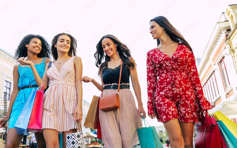 Sheer perfection. Low-angled photo of four female friends dressed in bright outfits, looking down and smiling while carrying several shopping bags. Sheer perfection. Low-angled photo of four female friends dressed in bright outfits, looking down and smiling while carrying several shopping bags.